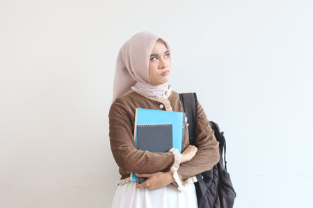 A young Muslim woman student thoughtfully holds her notebooks and backpack against a white wall. She is wearing a hijab and brown cardigan.の写真素材