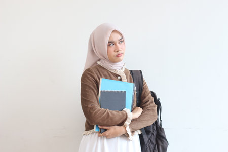 A young Muslim female student stands thoughtfully against a white wall, holding her books and backpack.の写真素材