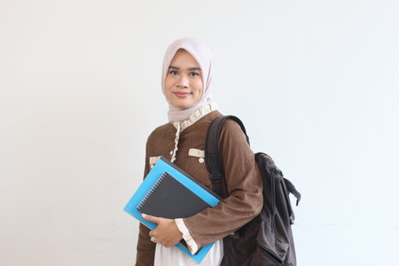 A cheerful young Muslim woman student smiles confidently while holding her books and backpack.  She is dressed in a brown jacket and headscarf.の写真素材