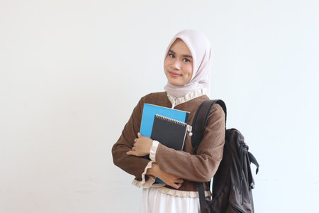 A young Muslim woman smiles confidently, holding her books and backpack ready for a day of learning.の写真素材