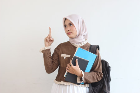 A young Muslim woman in a brown outfit holds notebooks and a backpack while thinking and pointing upward.  She is against a white background.の写真素材