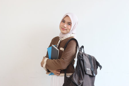 A cheerful young Muslim student girl smiles while holding books and a backpack. She looks confident and ready for school.の写真素材