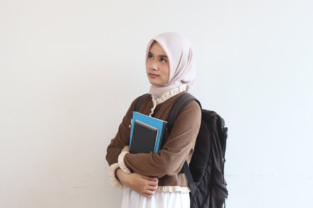 A young Muslim woman, wearing a hijab, stands thoughtfully, holding textbooks and a backpack.の写真素材