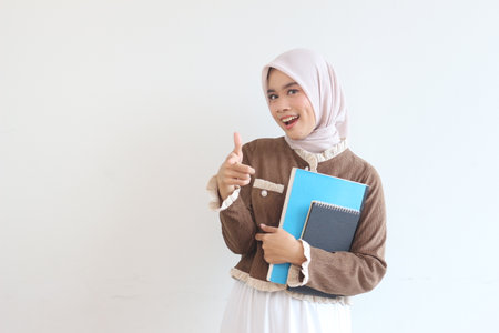 A cheerful young Muslim woman student in a hijab holding notebooks and pointing. She exudes happiness and readiness to learn.の写真素材