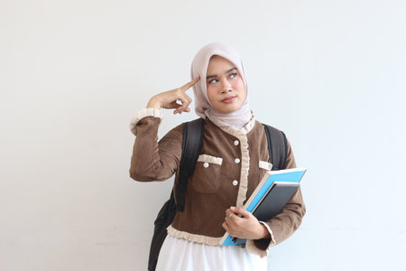 A young Muslim woman student thoughtfully holds books while wearing a hijab and backpack against a white wall.の写真素材