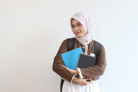 A cheerful young Muslim student girl smiles while holding books and wearing a backpack against a white backdrop.の写真素材