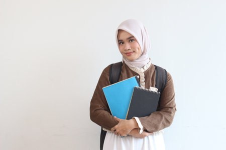 A cheerful young Muslim student girl in a hijab holds books and a backpack. She smiles confidently.の写真素材