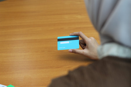 A woman's hand gently holds a light blue credit card against a light brown wooden background. The...