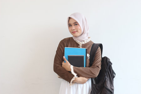 A young smiling Muslim woman in a hijab holds books and a backpack, suggesting a student on her way to or from classes.の写真素材