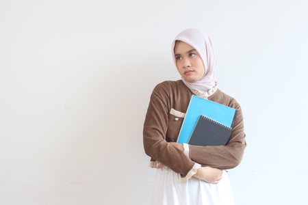 A young woman in a hijab holds notebooks, appearing thoughtful against a white backdrop.  The image conveys contemplation.の写真素材