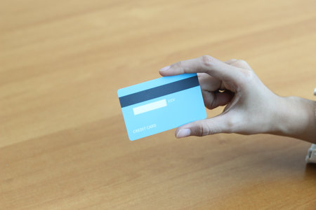 A person's hand holds a light blue credit card above a wooden table.  Represents online shopping and financial transactions.の写真素材
