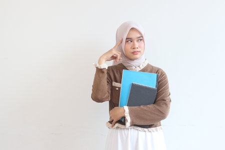 A young Muslim woman in a hijab is seen holding notebooks, deep in thought, exhibiting a contemplative mood.  The image is simple, emphasizing her intellectual expression.の写真素材