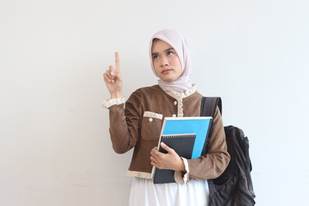 A young Muslim woman in hijab holds notebooks, backpack, thoughtfully pointing upwards.  She's contemplating an idea.の写真素材