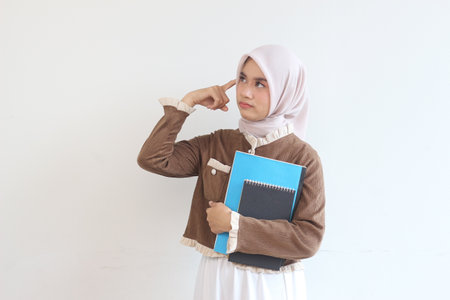 A young Muslim woman in a hijab holds notebooks, deep in thought.  She appears thoughtful and focused on a problem.の写真素材