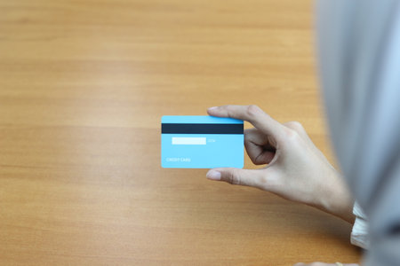 A hand holds a light blue credit card above a wooden table.  Focus is on the card and the hand.の写真素材