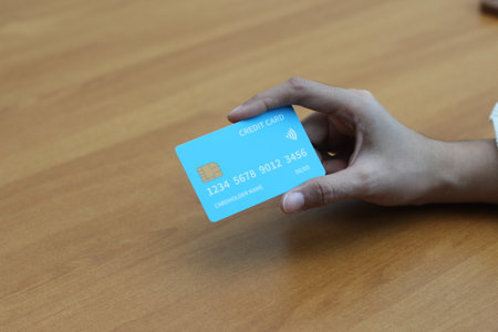 Close-up of a hand holding a light blue card on a wooden table. Represents online payment and financial technology.の写真素材