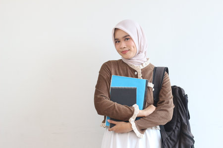 A cheerful young Muslim student smiles while holding her books and backpack against a simple white backgroundの写真素材