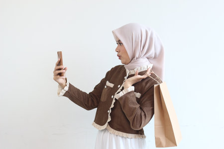 A young woman in a brown jacket and beige hijab takes a selfie with her phone while carrying a shopping bag.  She looks happy and content.の写真素材