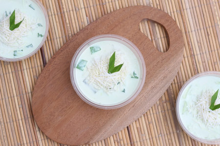 Three bowls of pandan flavored creamy dessert, topped with shredded cheese and pandan leaves. Served on a wooden board.の写真素材