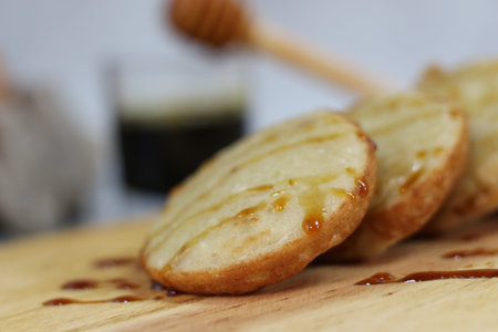 Close-up of three mini pancakes drizzled with honey on a wooden board. A blurry background shows a...