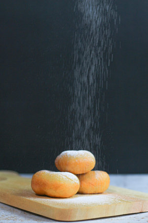 Three golden-brown donuts stacked on a wooden board, with powdered sugar falling on them.の写真素材