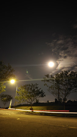 Nighttime scene showing a bright full moon illuminating the dark sky with clouds, trees silhouetted, and streetlights casting warm glow creating a peaceful atmosphere, ideal for nature and outdoor lighting marketing.の写真素材