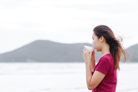the women  drink coffee near the beach. she stand on the beach and wear red shirtの写真素材