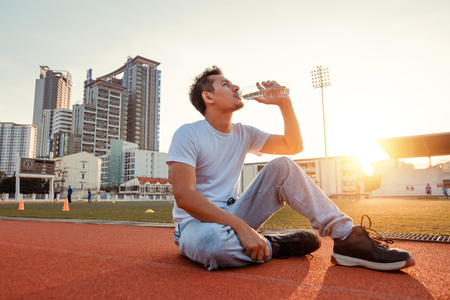 Caucasian man drinking water after exercises. Sport man sitting on track race at sport stadium and sunset.の写真素材