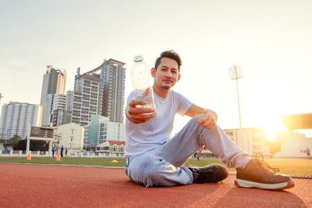 Caucasian man show water after exercises. Sport man sitting on track race at sport stadium and sunset.の写真素材