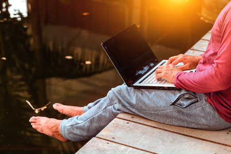 man working on laptop while sitting on wooden dock. legs in river waterの写真素材