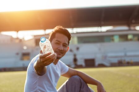 Caucasian man show water after exercises. Sport man sitting on track race at sport stadium and sunset.の写真素材