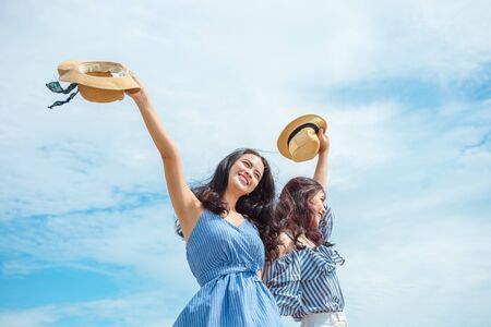 Two happy girl hold hat and raise arms in the sky. People traveling relaxing and enjoying  at summer.の写真素材