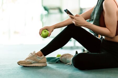 A girl hold green apple using smartphone in fitness gym. Woman exercise in gym fitness breaking and relax.の写真素材