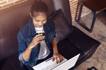 Young woman sitting on sofa at coffee shop and  drinking coffee while working on laptop.の写真素材