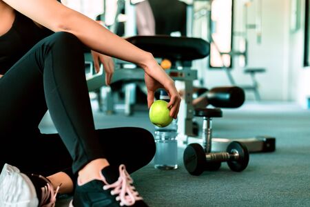 Woman exercise in gym fitness breaking and relax. hand  holding apple fruit after training sport and dumbbell, water bottle on the ground.の写真素材