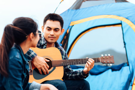 A man plays a guitar for a woman while camping beside the lake.