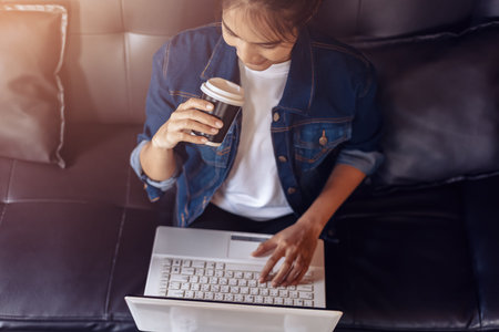Young woman sitting on sofa at coffee shop and  drinking coffee while working on laptop.