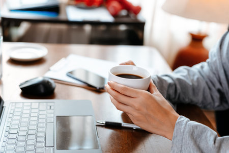 woman working on laptop and hold cup of coffee while sitting at working place.