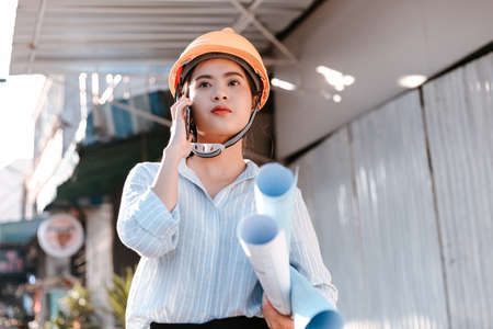 architect woman use mobile phone call with enginner or project manager between survey and checking at site construction. Architect woman hold blueprint.の写真素材