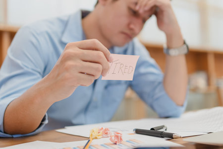 Businessman headache and hold sticky note with text TIRED. Tired, overworking after long time...