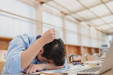 handsome businessman sleepy with coffee before working with laptop at desk in office. Working late night and start working early in the morning.の写真素材