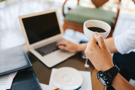 businessman holding coffee cup in hands and computer on table in coffee shop. Young freelancer drink coffee and working.の写真素材