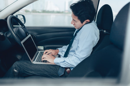 Businessman working in laptop and talking on the phone  in the drivers seat in his car.