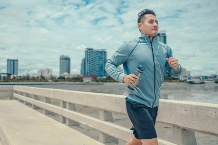 Young sport man hold water bottle and wearing earphone while jogging and running on the road and city backgound.の写真素材