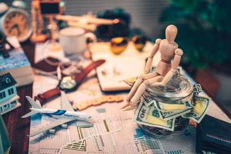 Wood human doll sitting on Saving money in glass jar for travel, holiday, vocation on road map on the table. Travel budget concept.の写真素材