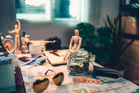 Wood human doll sitting on Saving money in glass jar for travel, holiday, vocation on road map on the table. Travel budget concept.の写真素材