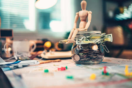Wood human doll sitting on Saving money in glass jar for travel, holiday, vocation on road map on the table. Travel budget concept.の写真素材