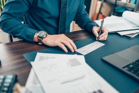 Business man hand writing and signing white blank bank check book and dollar bill, coin, laptop and...