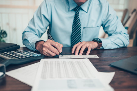 Business man hand writing and signing white blank bank check book and dollar bill, coin, laptop and graph chart on the desk at office. Payment by check, paycheck, payroll concept.の写真素材