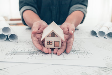 Architect engineer hands hold and protect mini house model on bueprint. Protect House, cost planning design and construction concept.の写真素材